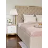 Beżowe łóżko glamour SPECTRE z pikowanym zagłówkiem, obok biała szafka nocna i elegancka lampa.