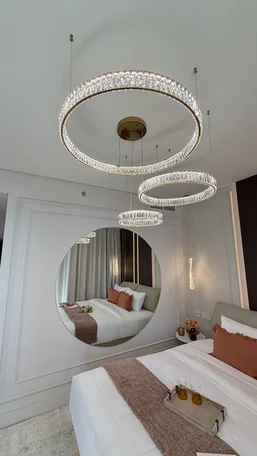 Trzy kryształowe ringi żyrandola Crystal XXL nad łóżkiem widziane z rogu jasnej sypialni z okrągłym lustrem.
