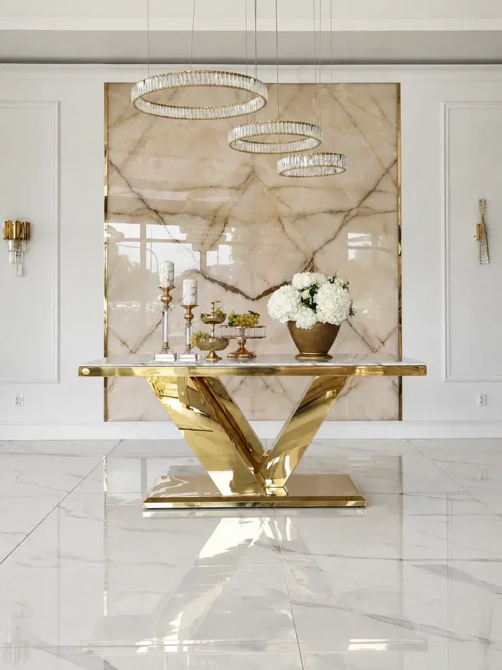 Stół glamour LV Collection z białym blatem i złotą podstawą, ustawiony centralnie w jasnym, przestronnym wnętrzu z marmurową ścianą i wiszącymi lampami.