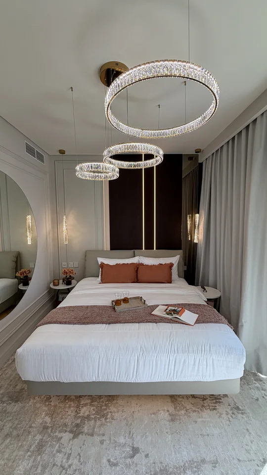 Potrójny żyrandol Crystal XXL w złocie nad łóżkiem w eleganckiej sypialni z jasnymi zasłonami i ciemną ścianą.
