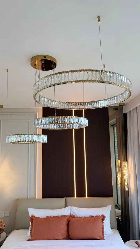 Kryształowe ringi żyrandola Crystal XXL w złotej oprawie zawieszone nad łóżkiem z poduszkami w kolorze terakoty.